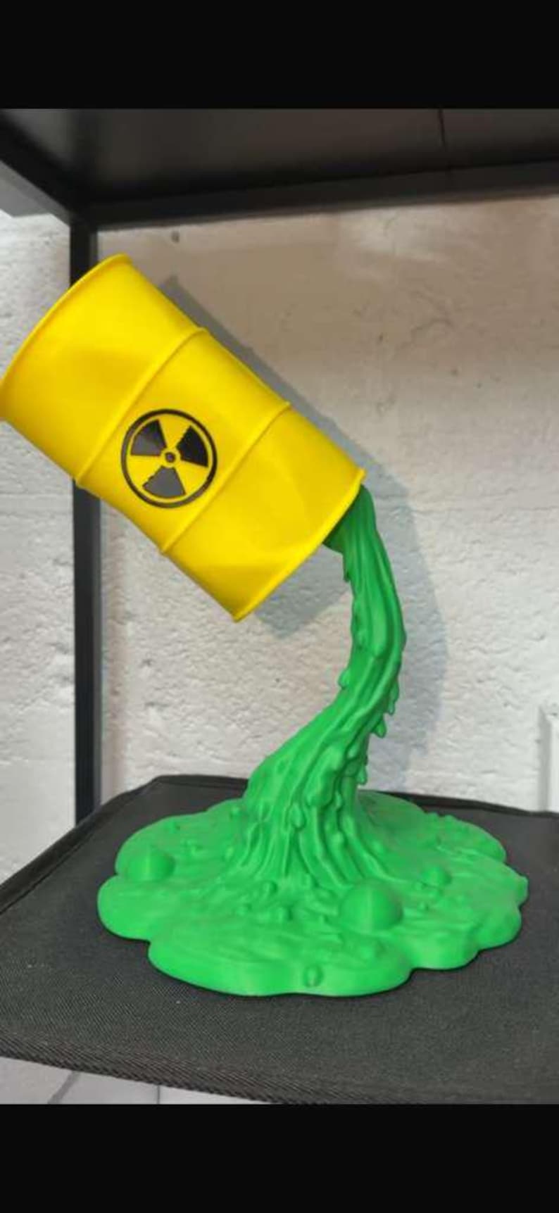 May include: A yellow barrel with a black and white radiation symbol is tilted, with green liquid pouring out. The liquid forms a puddle at the base. The sculpture is a unique decorative item.