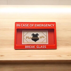 May include: A red break glass box with the words "IN CASE OF EMERGENCY" and "BREAK GLASS" printed on it. The box contains a white card with the words "Platform 9 3/4" and a black and white image of the Hogwarts crest.