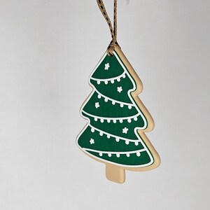 May include: A Christmas tree-shaped ornament with a green design and white accents. The ornament is made to look like a cookie, with a tan border and a brown ribbon for hanging. The tree has white string lights and star details.