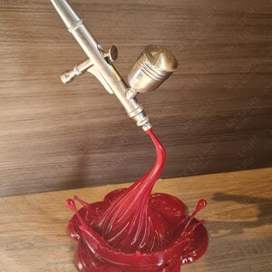 May include: A silver airbrush model with a red paint splash base. The airbrush is angled downward, with a stream of red paint flowing into a stylized splash. The base is a glossy, flower-like shape.