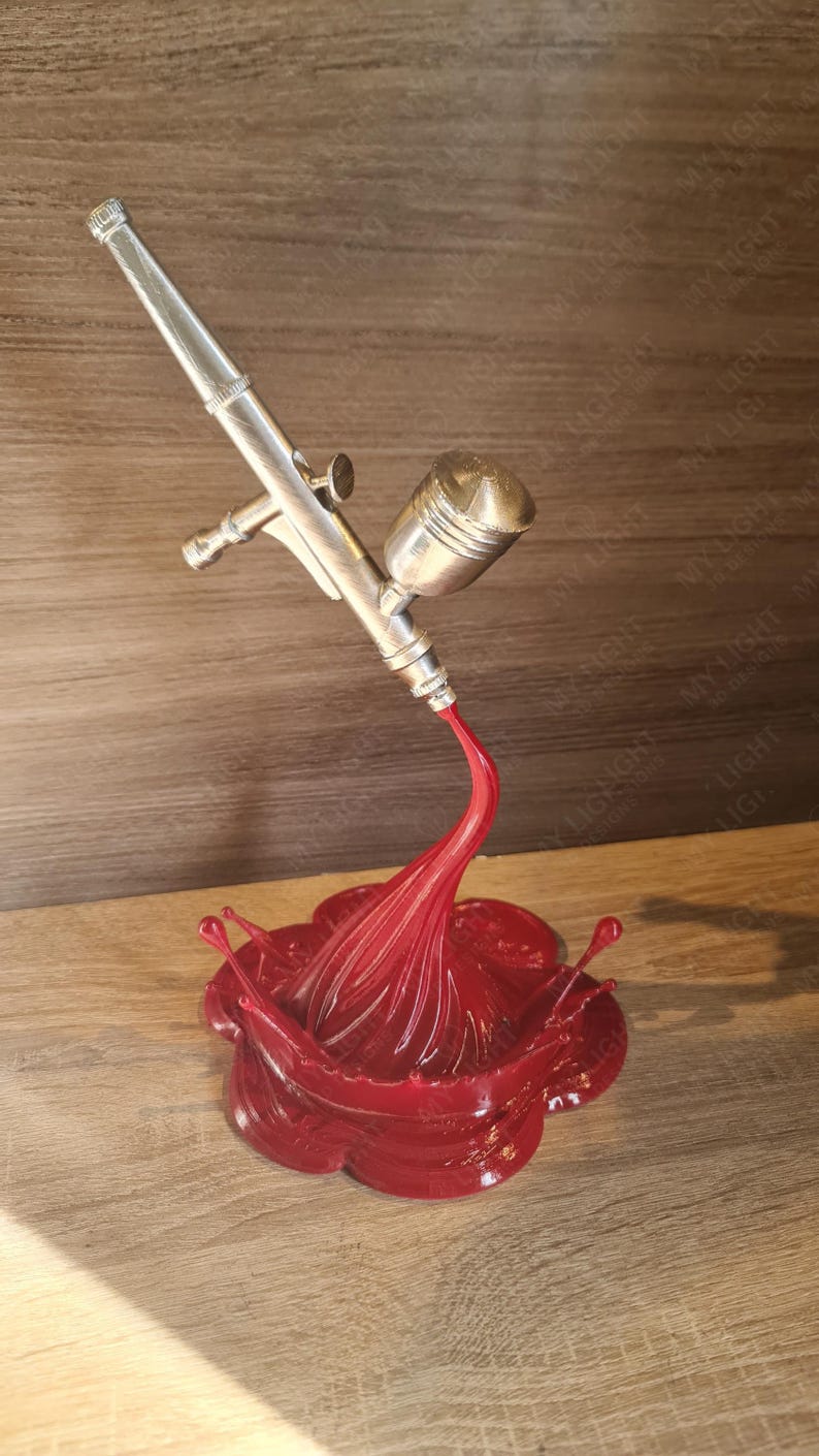 May include: A silver airbrush model with a red paint splash base. The airbrush is angled downward, with a stream of red paint flowing into a stylized splash. The base is a glossy, flower-like shape.