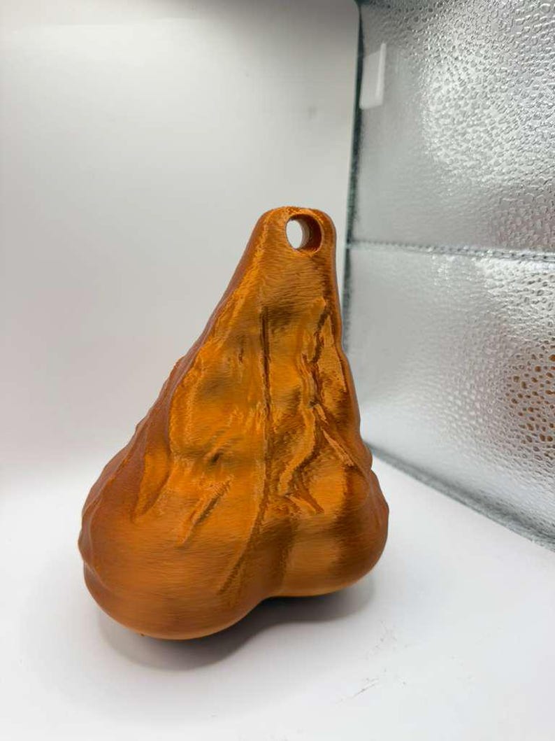 May include: A metallic copper-colored, three-dimensional object with a textured, organic, and irregular shape. The object features a prominent, rounded base that tapers upward to a narrow point, which contains a single, circular hole for hanging.