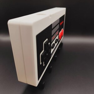May include: A white and black retro video game controller design. The controller has a classic button layout with red, grey, and black accents. The design is reminiscent of a vintage gaming console.