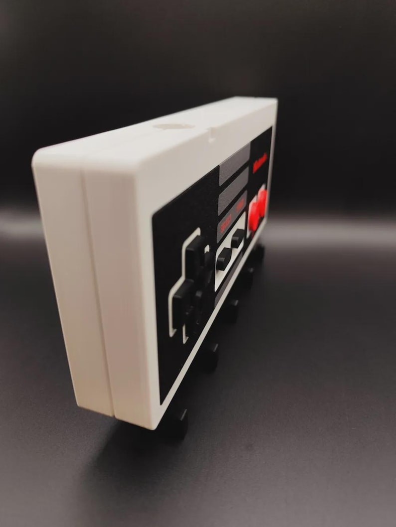 May include: A white and black retro video game controller design. The controller has a classic button layout with red, grey, and black accents. The design is reminiscent of a vintage gaming console.
