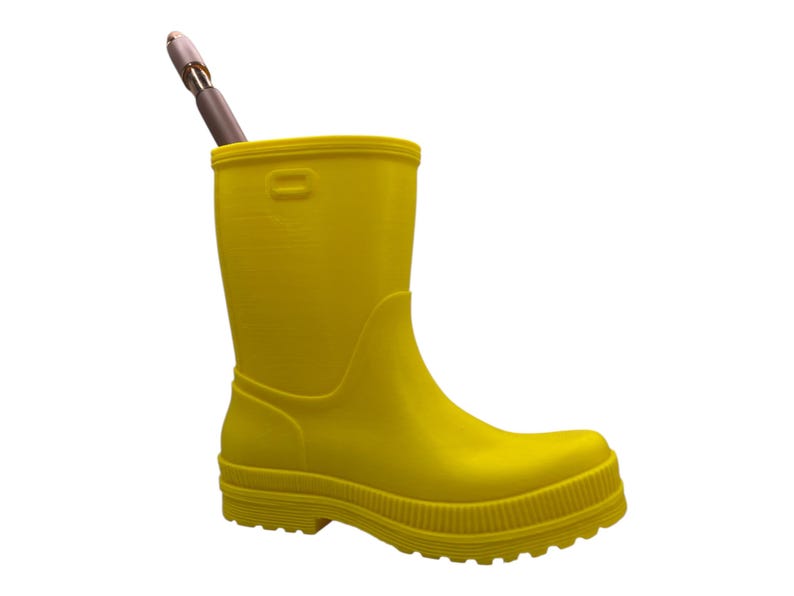 May include: A bright yellow rubber boot, possibly a pencil holder, with a brown and silver pen inside. The boot has a ridged sole and a smooth, glossy finish. The background is plain white.