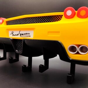 Puede incluir: Un llavero de pared amarillo y negro con forma de parte trasera de coche deportivo. El llavero tiene una rejilla negra, tubos de escape y luces traseras rojas. El nombre "Suno Ferrari" est&aacute; impreso en la matr&iacute;cula.
