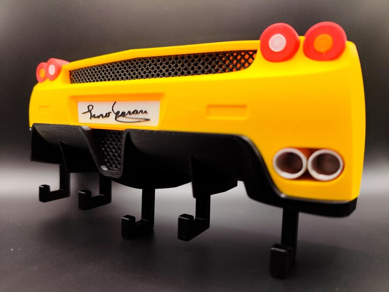 Puede incluir: Un llavero de pared amarillo y negro con forma de parte trasera de coche deportivo. El llavero tiene una rejilla negra, tubos de escape y luces traseras rojas. El nombre "Suno Ferrari" est&aacute; impreso en la matr&iacute;cula.