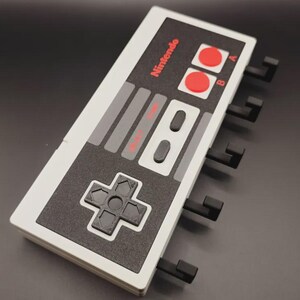 May include: A wall-mounted organiser shaped like a classic Nintendo controller. The design features a white and grey colour scheme with red buttons and the Nintendo logo. It has several black hooks for hanging items.