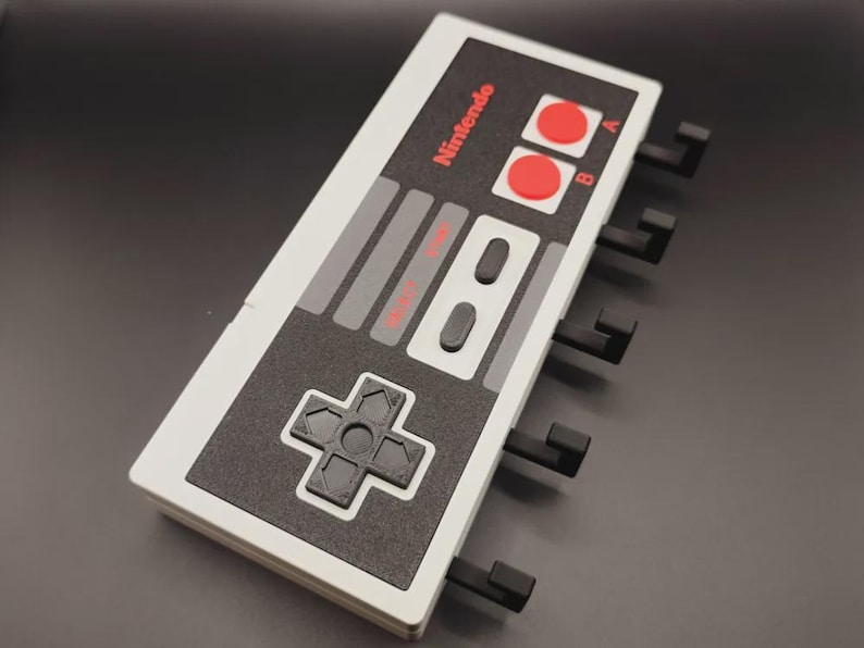 May include: A wall-mounted organiser shaped like a classic Nintendo controller. The design features a white and grey colour scheme with red buttons and the Nintendo logo. It has several black hooks for hanging items.