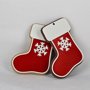 May include: Two Christmas stocking ornaments. The stockings are red with white tops and a white snowflake design. One stocking has a brown border, and the other has a tan border. Both have a hole for hanging.