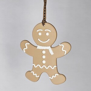 K&ouml;nnte beinhalten: Ein Lebkuchenmann-Ornament mit braunem K&ouml;rper, wei&szlig;en Zuckergussdetails und einem wei&szlig;en Schal. Das Ornament hat ein l&auml;chelndes Gesicht und wird von einem braunen Band gehalten. Eine festliche Dekoration f&uuml;r die Weihnachtszeit.