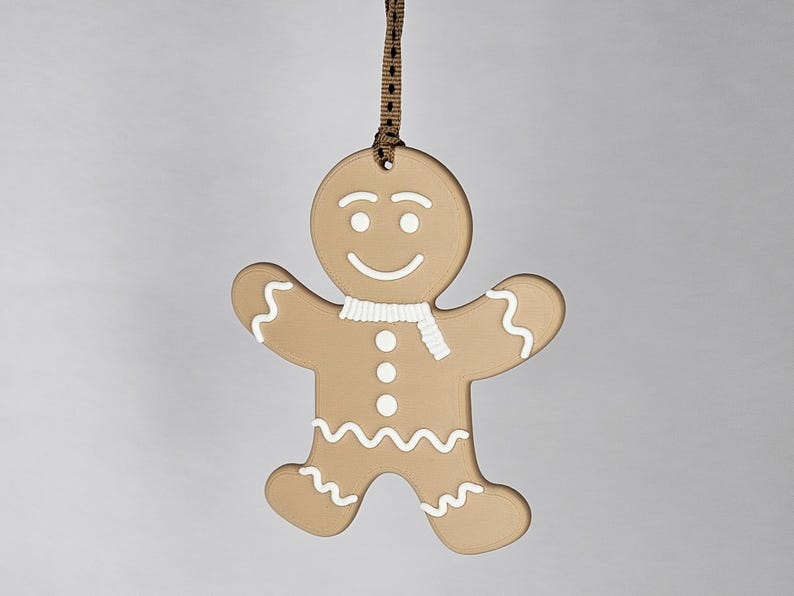 K&ouml;nnte beinhalten: Ein Lebkuchenmann-Ornament mit braunem K&ouml;rper, wei&szlig;en Zuckergussdetails und einem wei&szlig;en Schal. Das Ornament hat ein l&auml;chelndes Gesicht und wird von einem braunen Band gehalten. Eine festliche Dekoration f&uuml;r die Weihnachtszeit.