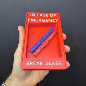 May include: A rectangular red plastic frame with the white text IN CASE OF EMERGENCY and BREAK GLASS. Inside the recessed center, two crayons are positioned diagonally, one blue and one red, each with the word CRAYONS printed on them.