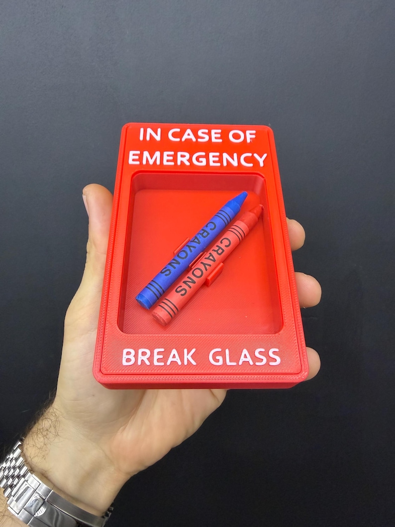 May include: A rectangular red plastic frame with the white text IN CASE OF EMERGENCY and BREAK GLASS. Inside the recessed center, two crayons are positioned diagonally, one blue and one red, each with the word CRAYONS printed on them.