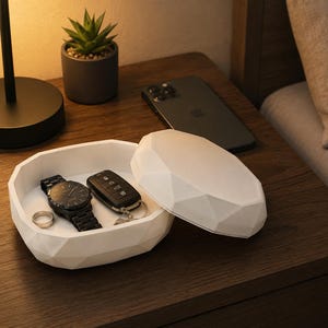 Puede incluir: Un organizador de mesita de noche blanco, de forma geom&eacute;trica, con tapa, que contiene un reloj negro, llaves de coche y un anillo. Un smartphone negro descansa sobre la mesita de noche de madera, junto a una peque&ntilde;a planta en maceta y una l&aacute;mpara.