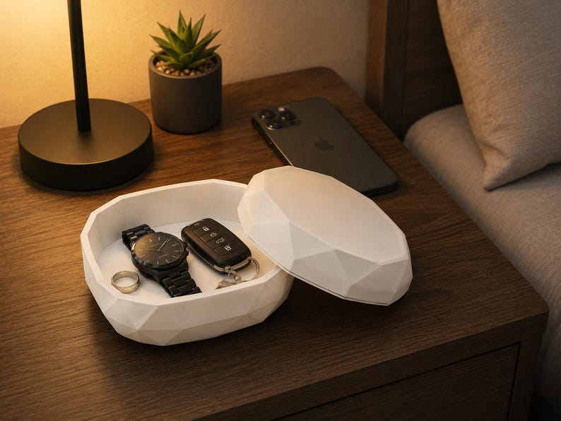 Puede incluir: Un organizador de mesita de noche blanco, de forma geom&eacute;trica, con tapa, que contiene un reloj negro, llaves de coche y un anillo. Un smartphone negro descansa sobre la mesita de noche de madera, junto a una peque&ntilde;a planta en maceta y una l&aacute;mpara.