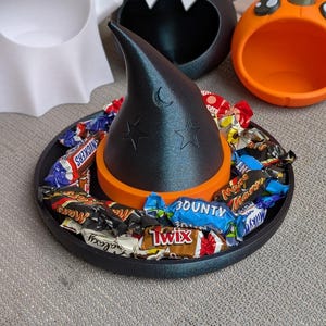 May include: A black witch hat candy dish with a crescent moon and star design, filled with wrapped sweets. The hat sits on a black tray with an orange base. Other Halloween-themed containers are in the background.