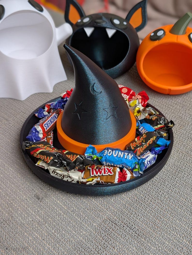 May include: A black witch hat candy dish with a crescent moon and star design, filled with wrapped sweets. The hat sits on a black tray with an orange base. Other Halloween-themed containers are in the background.
