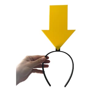 May include: A yellow arrow-shaped headpiece attached to a black headband. The arrow points downward. The headband is held by a hand against a white background. The arrow is a solid, bright yellow color.