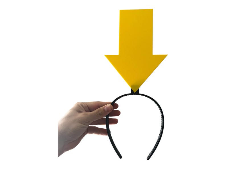 May include: A yellow arrow-shaped headpiece attached to a black headband. The arrow points downward. The headband is held by a hand against a white background. The arrow is a solid, bright yellow color.