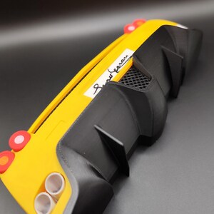 Puede incluir: Un modelo de coche amarillo y negro con aler&oacute;n trasero. El coche tiene dos tubos de escape, luces traseras rojas y una etiqueta blanca con texto en cursiva. El modelo es una r&eacute;plica en miniatura de un coche deportivo.