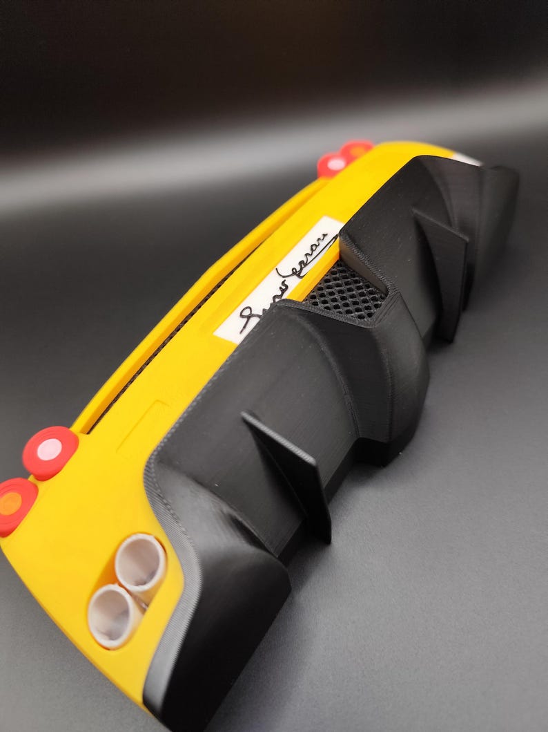 Puede incluir: Un modelo de coche amarillo y negro con aler&oacute;n trasero. El coche tiene dos tubos de escape, luces traseras rojas y una etiqueta blanca con texto en cursiva. El modelo es una r&eacute;plica en miniatura de un coche deportivo.