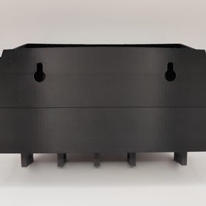 May include: A black, wall-mounted organizer with a decorative top edge. It features two keyhole slots for hanging and several small, rectangular supports along the bottom. The organizer is made of a solid, matte material.