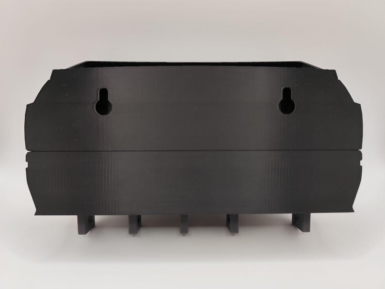 May include: A black, wall-mounted organizer with a decorative top edge. It features two keyhole slots for hanging and several small, rectangular supports along the bottom. The organizer is made of a solid, matte material.