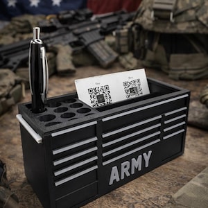 May include: Black desk organizer with the word "ARMY" in white. It has a pen holder, a slot for business cards, and a small drawer. A pen is in the pen holder. The background includes an American flag and military equipment.