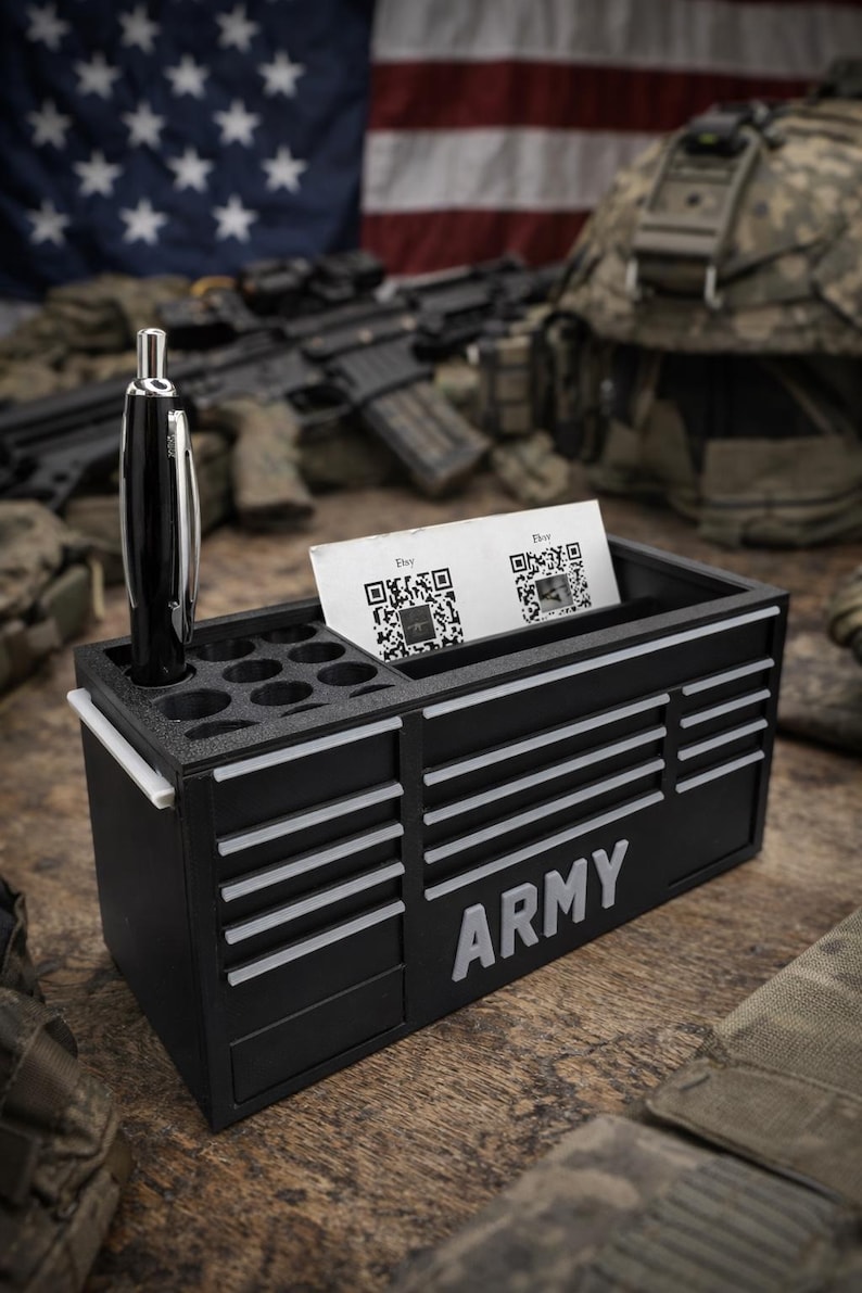 May include: Black desk organizer with the word "ARMY" in white. It has a pen holder, a slot for business cards, and a small drawer. A pen is in the pen holder. The background includes an American flag and military equipment.