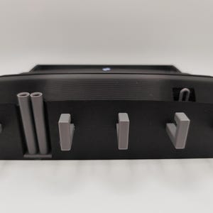 May include: A black, rectangular wall-mounted key holder with gray hooks and cylindrical accents. The key holder has a textured surface and a small, blue light on top. The design is modern and minimalist.