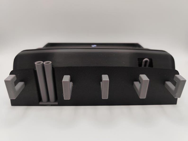 May include: A black, rectangular wall-mounted key holder with gray hooks and cylindrical accents. The key holder has a textured surface and a small, blue light on top. The design is modern and minimalist.
