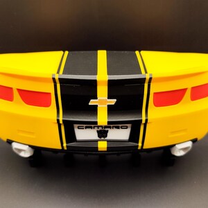 Puede incluir: Un coche de juguete amarillo con rayas negras en el centro. El coche tiene luces traseras rojas, un emblema Chevrolet plateado y la palabra "CAMARO" en la matr&iacute;cula. El coche tiene tubos de escape blancos.