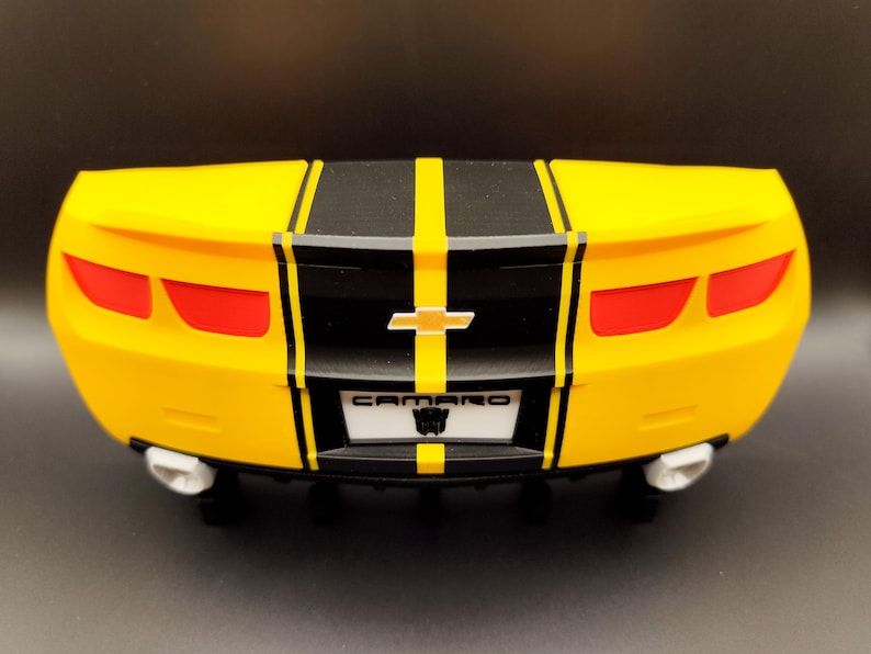 Puede incluir: Un coche de juguete amarillo con rayas negras en el centro. El coche tiene luces traseras rojas, un emblema Chevrolet plateado y la palabra "CAMARO" en la matr&iacute;cula. El coche tiene tubos de escape blancos.