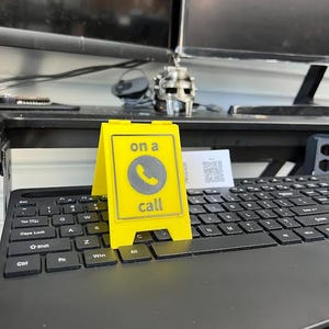 May include: A bright yellow desk sign with the words "on a call" and a phone icon. The sign is placed on a black keyboard. The sign is a-frame style and is made of a solid material.