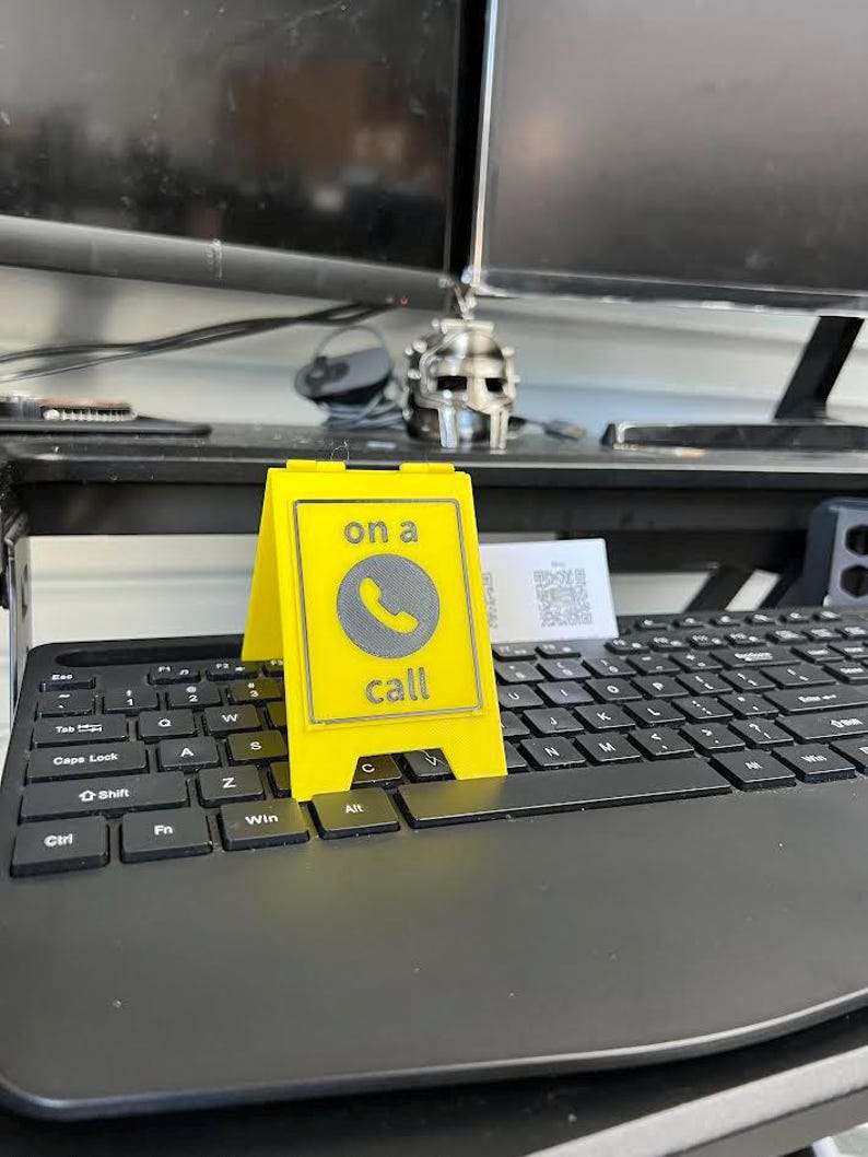 May include: A bright yellow desk sign with the words "on a call" and a phone icon. The sign is placed on a black keyboard. The sign is a-frame style and is made of a solid material.