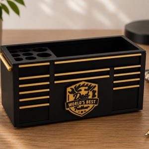 May include: A black desk organizer shaped like a toolbox, with gold accents and the text "Dad World's Best Father." It features compartments for pens and tools, and gold handles. A stylish and functional gift.