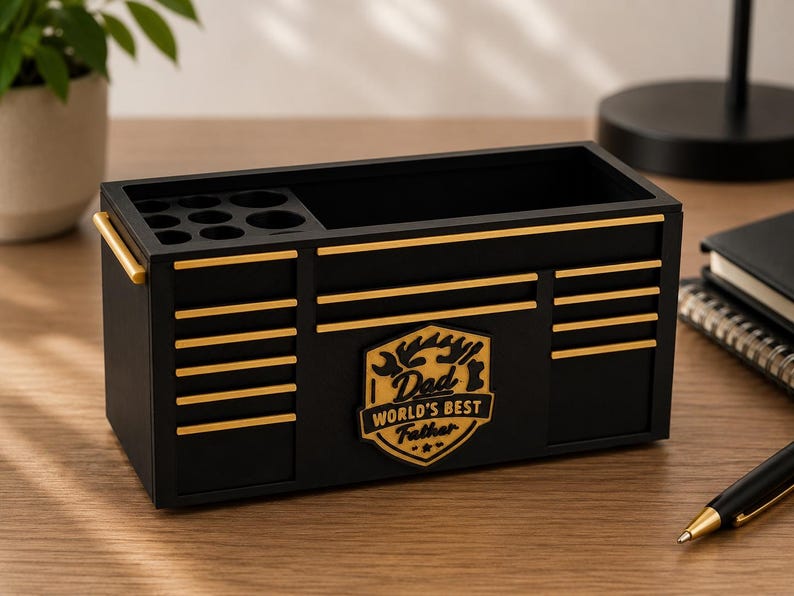 May include: A black desk organizer shaped like a toolbox, with gold accents and the text "Dad World's Best Father." It features compartments for pens and tools, and gold handles. A stylish and functional gift.