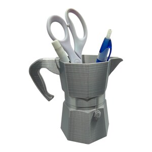 May include: A gray, 3D-printed desk organizer shaped like a stovetop espresso maker. It holds a pair of white scissors, a white marker, and a blue pen. The organizer has a handle and a spout, and is designed to hold office supplies.