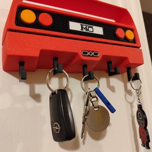 May include: A red key holder shaped like a car's rear end, with the text "F40" on a white rectangle. It has four black hooks for keys, with several keys hanging from them. The key holder is mounted on a white wall.