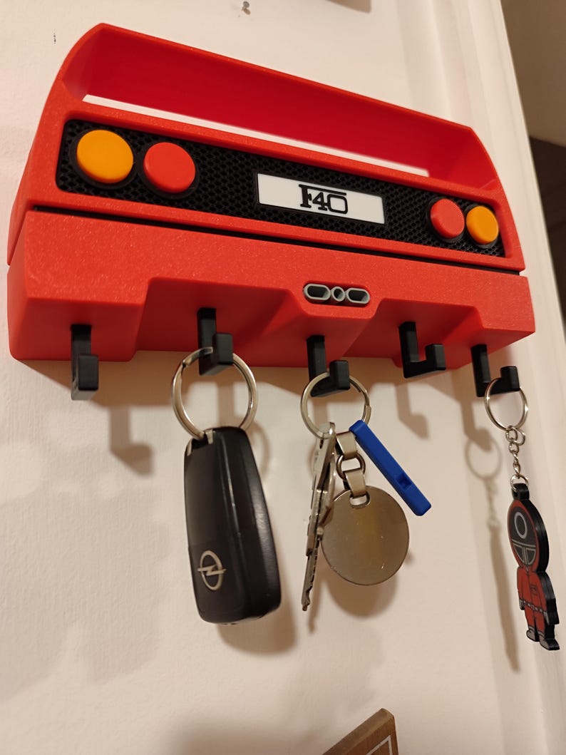 May include: A red key holder shaped like a car's rear end, with the text "F40" on a white rectangle. It has four black hooks for keys, with several keys hanging from them. The key holder is mounted on a white wall.