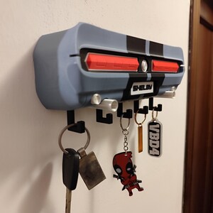 K&ouml;nnte beinhalten: Ein an der Wand befestigter Schl&uuml;sselhalter in Form des Hecks eines klassischen Autos, in Grau- und Schwarzt&ouml;nen, mit roten R&uuml;ckleuchten. Der Schl&uuml;sselhalter hat mehrere Haken f&uuml;r Schl&uuml;ssel und das Wort "Shelby" ist sichtbar. Mehrere Schl&uuml;ssel und Schl&uuml;sselanh&auml;nger h&auml;ngen an den Haken.