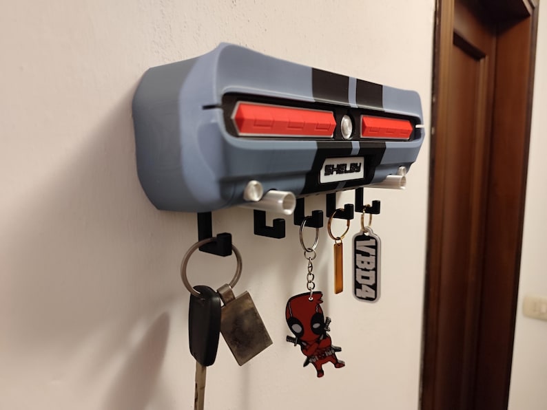 K&ouml;nnte beinhalten: Ein an der Wand befestigter Schl&uuml;sselhalter in Form des Hecks eines klassischen Autos, in Grau- und Schwarzt&ouml;nen, mit roten R&uuml;ckleuchten. Der Schl&uuml;sselhalter hat mehrere Haken f&uuml;r Schl&uuml;ssel und das Wort "Shelby" ist sichtbar. Mehrere Schl&uuml;ssel und Schl&uuml;sselanh&auml;nger h&auml;ngen an den Haken.