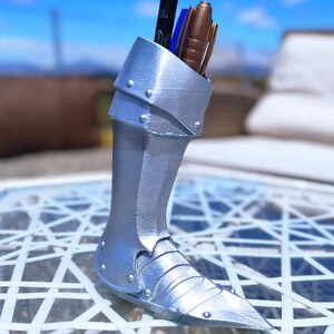May include: A silver-coloured pen holder in the shape of a knight's greave, with a pointed toe and riveted details. The container holds several pens and pencils. The pen holder is made of a metallic material and sits on a white patterned surface.