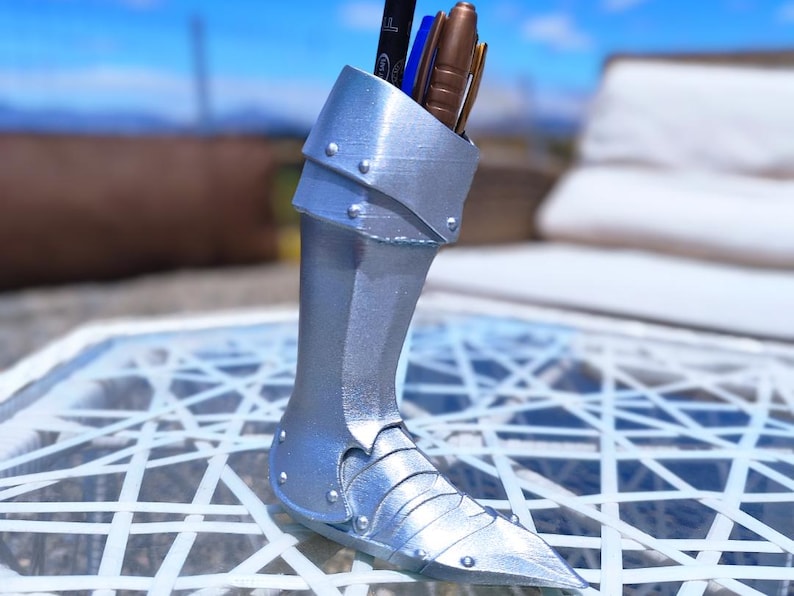 May include: A silver-coloured pen holder in the shape of a knight's greave, with a pointed toe and riveted details. The container holds several pens and pencils. The pen holder is made of a metallic material and sits on a white patterned surface.