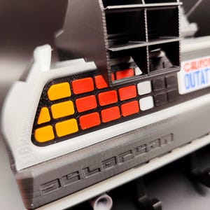 Pode incluir: Modelo detalhado da sec&ccedil;&atilde;o traseira de um carro DeLorean, com uma carro&ccedil;aria cinzenta com a palavra "DELOREAN" em relevo. As luzes traseiras t&ecirc;m luzes quadradas laranja, vermelhas e brancas. Uma estrutura de ventila&ccedil;&atilde;o preta est&aacute; posicionada acima do carro. A matr&iacute;cula diz "CALIFORNIA OUTATIME".