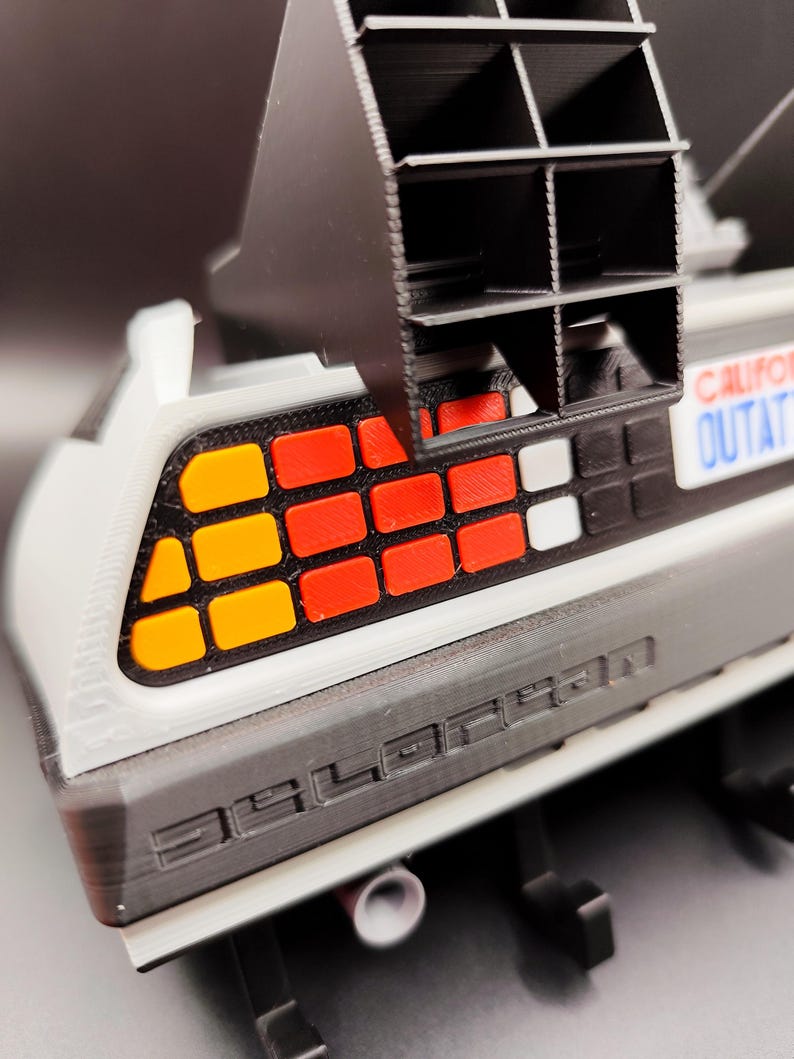 Pode incluir: Modelo detalhado da sec&ccedil;&atilde;o traseira de um carro DeLorean, com uma carro&ccedil;aria cinzenta com a palavra "DELOREAN" em relevo. As luzes traseiras t&ecirc;m luzes quadradas laranja, vermelhas e brancas. Uma estrutura de ventila&ccedil;&atilde;o preta est&aacute; posicionada acima do carro. A matr&iacute;cula diz "CALIFORNIA OUTATIME".