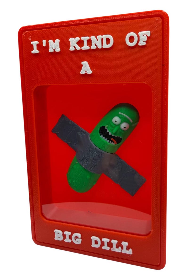 May include: A red framed display featuring a green pickle character with a gray weapon. The text "I'M KIND OF A" is at the top, and "BIG DILL" is at the bottom in white letters. The pickle has a smiling face.