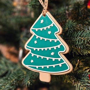 K&ouml;nnte beinhalten: Ein weihnachtsbaumf&ouml;rmiger Ornament mit t&uuml;rkisgr&uuml;ner Innenseite und wei&szlig;en Zuckergussdetails. Der Anh&auml;nger h&auml;ngt an einem braunen Band und vor einem Weihnachtsbaum.