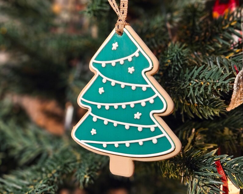 K&ouml;nnte beinhalten: Ein weihnachtsbaumf&ouml;rmiger Ornament mit t&uuml;rkisgr&uuml;ner Innenseite und wei&szlig;en Zuckergussdetails. Der Anh&auml;nger h&auml;ngt an einem braunen Band und vor einem Weihnachtsbaum.